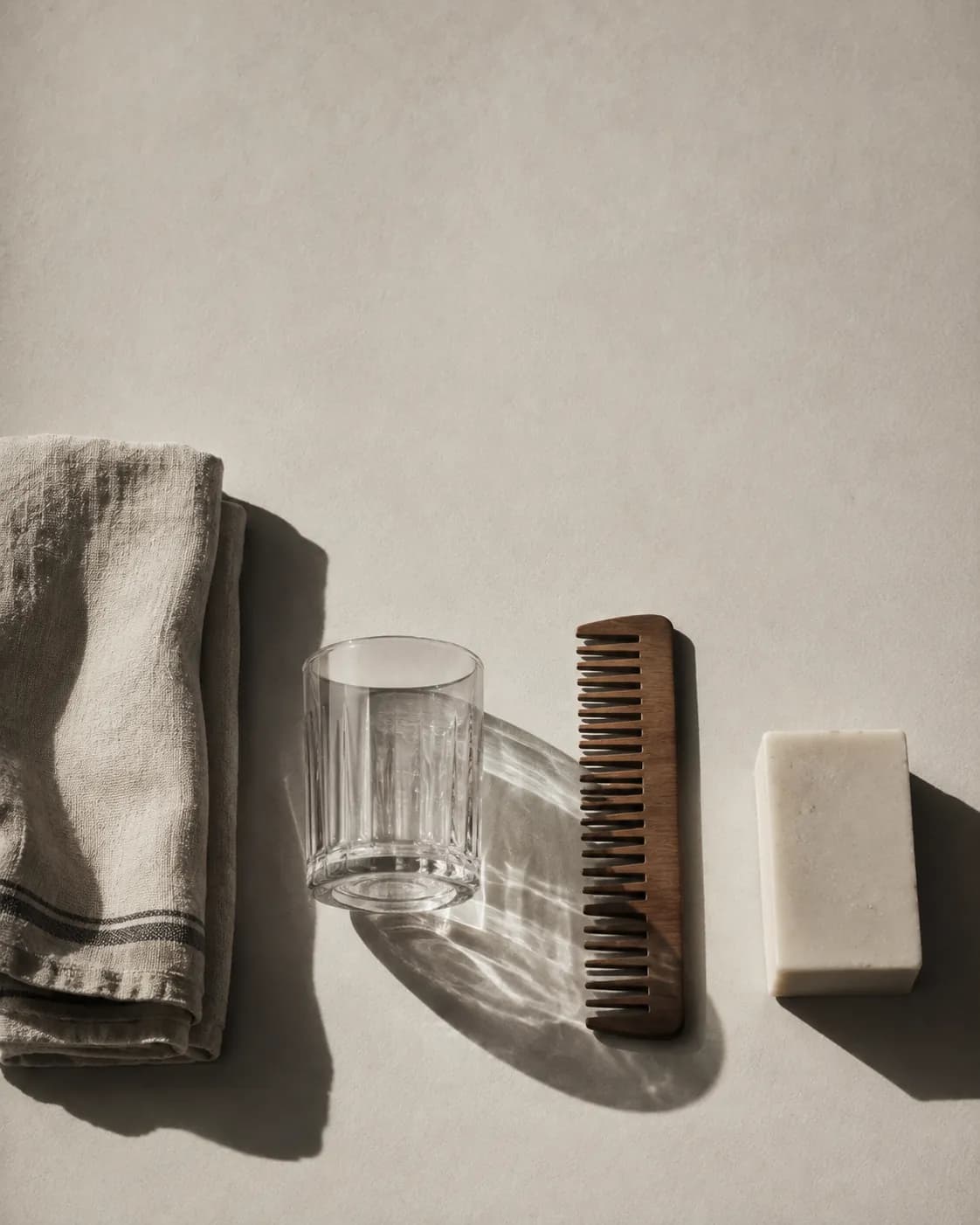 A folded linen towel, a glass tumbler, a wooden comb and a bar of soap laid out in a row on warm off-white paper.