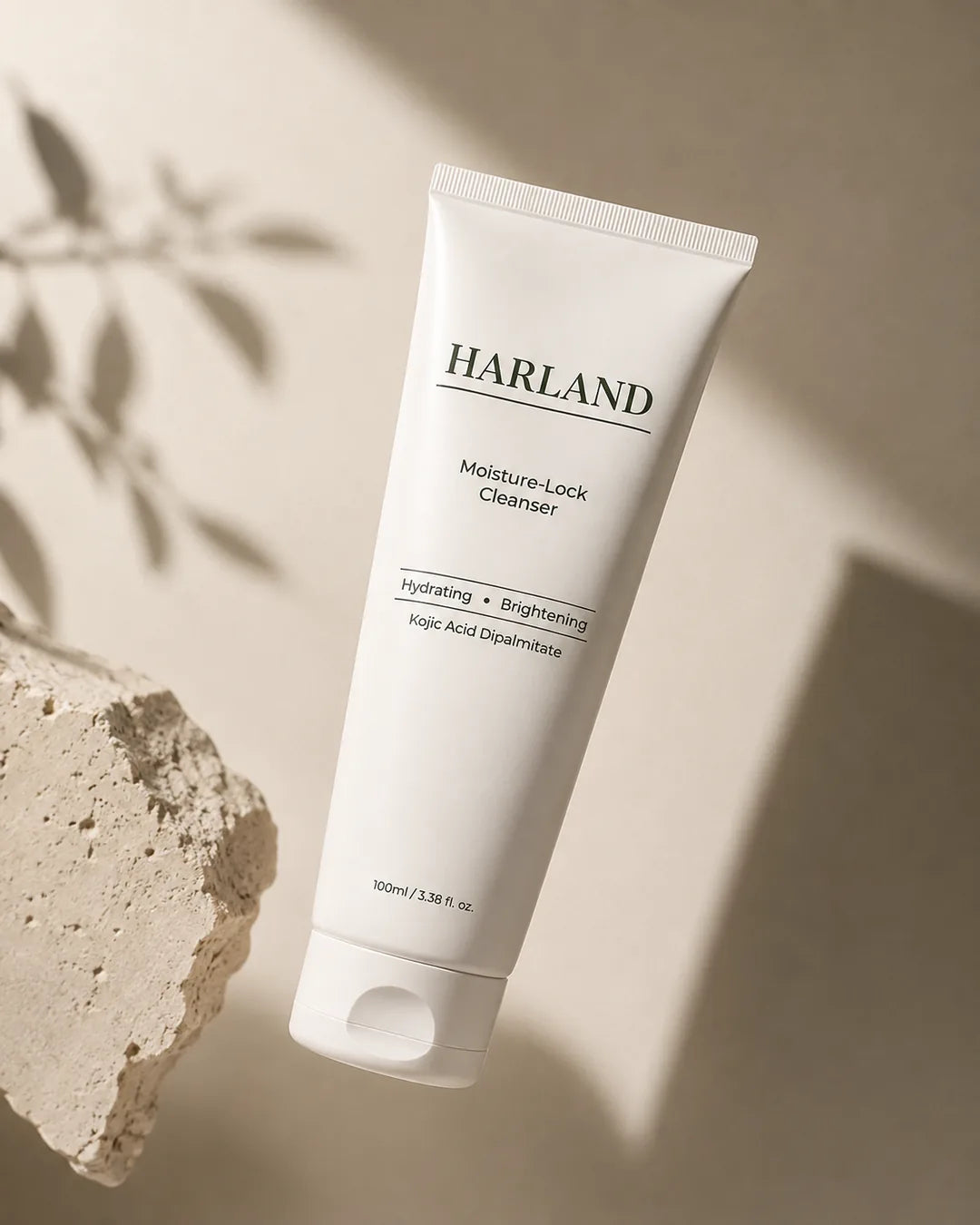 The HARLAND Moisture-Lock Cleanser, a matte cream 100 ml tube, propped against a block of travertine in daylight with leaf shadows falling across the wall behind it.